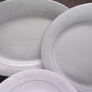 May include: A collection of white porcelain dinnerware with a delicate floral pattern and a silver trim. The set includes an oval platter and several round plates. The plates are arranged on a dark surface.