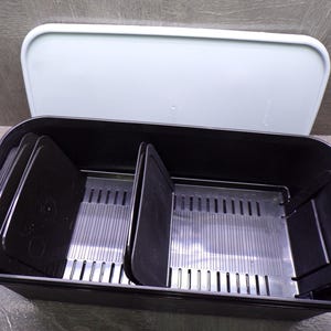 May include: A black plastic storage container with a light blue lid. The container has multiple compartments with removable dividers and a slotted tray. The lid is slightly ajar, revealing the interior. The container is designed for organizing and storing items.