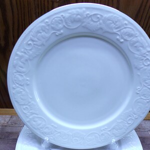 Pu&ograve; includere: Piatto in ceramica bianca con un motivo floreale e di vite in rilievo sul bordo. Il piatto ha un centro liscio e leggermente concavo. Il piatto &egrave; probabilmente per servire cibo. Il piatto &egrave; su un supporto di plastica trasparente.