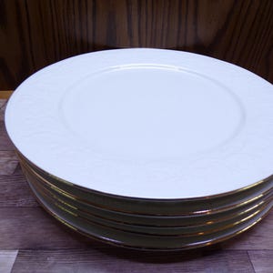 Pu&ograve; includere: Una pila di piatti in ceramica bianca con bordo dorato. I piatti hanno un sottile motivo floreale in rilievo sul bordo. I piatti sono impilati su una superficie di legno.
