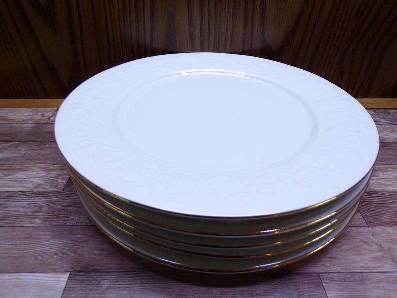 Pu&ograve; includere: Una pila di piatti in ceramica bianca con bordo dorato. I piatti hanno un sottile motivo floreale in rilievo sul bordo. I piatti sono impilati su una superficie di legno.