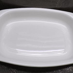 May include: A white, rectangular baking dish with rounded corners. The dish has a smooth, glossy surface and a slightly raised rim. The interior is a bright white, reflecting light. This dish is suitable for baking and serving.