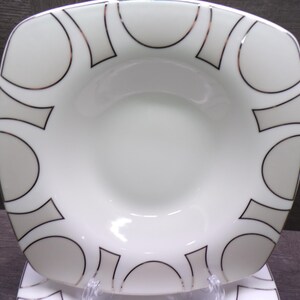 May include: White square-shaped bowl with a rounded interior. The rim features a repeating pattern of gray circles and rectangles outlined in black. The bowl is set on a clear stand, showcasing a modern design.