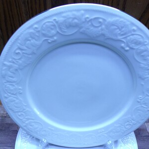 Pu&ograve; includere: Un piatto bianco di forma ovale con un motivo floreale e a foglie in rilievo sul bordo. Il piatto in ceramica &egrave; presentato su un supporto trasparente. Il design del piatto &egrave; elegante e adatto per servire cibo.