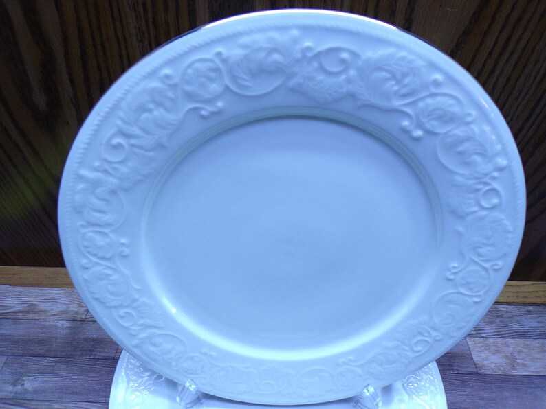Pu&ograve; includere: Un piatto bianco di forma ovale con un motivo floreale e a foglie in rilievo sul bordo. Il piatto in ceramica &egrave; presentato su un supporto trasparente. Il design del piatto &egrave; elegante e adatto per servire cibo.