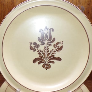 May include: Cream-colored ceramic plate with a brown floral design in the center. The plate has a brown border and a matching floral pattern. The plate is part of a dinnerware set.