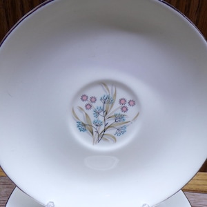May include: A white saucer with a delicate floral design in the center. The design features blue and pink flowers with green stems. The saucer has a silver rim and is part of a vintage dish set.