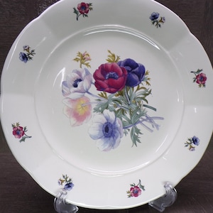 May include: White ceramic plate with a scalloped edge, decorated with a bouquet of anemone flowers in shades of purple, red, and white. Small floral accents are scattered around the rim. A decorative piece for serving.