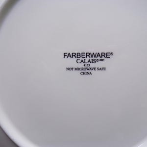 Pu&ograve; includere: Piatto bianco Farberware Calais con la scritta "FARBERWARE CALAIS &copy;2001 4173 NOT MICROWAVE SAFE CHINA" stampata in nero. Il piatto ha una superficie liscia e lucida.