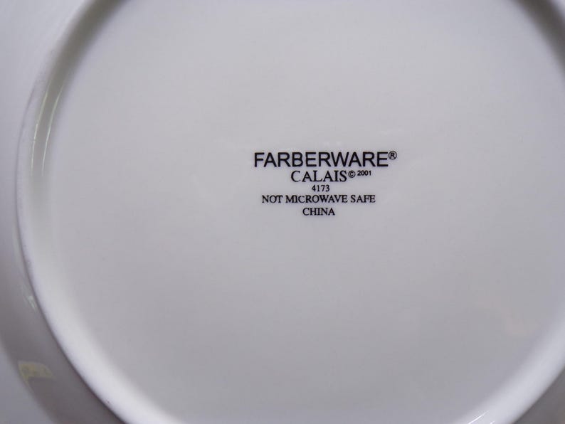 Pu&ograve; includere: Piatto bianco Farberware Calais con la scritta "FARBERWARE CALAIS &copy;2001 4173 NOT MICROWAVE SAFE CHINA" stampata in nero. Il piatto ha una superficie liscia e lucida.