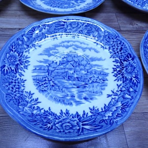 May include: Blue and white decorative plates with a floral and landscape design. The plates have a scalloped edge and a detailed illustration of a building and bridge. The plates are arranged on a wooden surface.