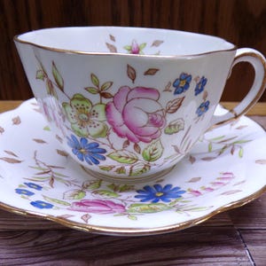 May include: A white teacup and saucer set with a floral design. The cup and saucer have a gold trim and are decorated with pink roses, blue and green flowers, and green leaves. The saucer has a scalloped edge.
