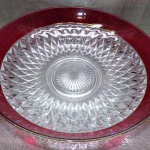 May include: A clear glass serving bowl with a ruby red rim. The bowl features a diamond-cut pattern on the interior and a circular design in the center. The rim has a gold trim.