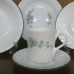 May include: A set of white ceramic dishes with a green ivy design. The set includes a teacup, saucer, and three plates.