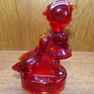 May include: A red glass figurine of a bird with a round base. The bird is sitting with its head up and wings spread. The glass is transparent, allowing light to pass through, and has a glossy finish. The figurine is a decorative item.