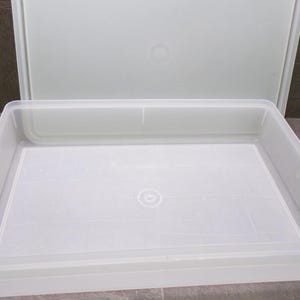 May include: A rectangular, semi-transparent plastic storage container with a low profile and a matching lid positioned upright behind it. The base of the container features a subtle grid pattern and a central circular indentation for structural support.