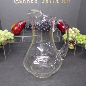 May include: A clear glass pitcher with a wide base and a handle. The pitcher is empty and has a slight curve to the body.