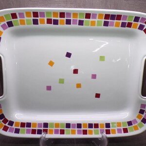 May include: A rectangular white ceramic serving tray with integrated side handles. The rim features a colorful mosaic-style border of small purple, orange, lime green, and maroon squares. The center is decorated with scattered squares in the same colors.
