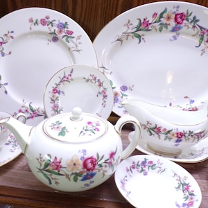 May include: A white porcelain tea set with floral designs. The set includes a teapot, plates, a gravy boat, and cups. The floral pattern features pink, purple, yellow, and blue flowers with green leaves, and gold trim.