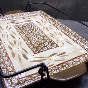 May include: A rectangular ceramic serving tray with a cream-colored center featuring a brown floral pattern, surrounded by a border of small white flowers on a brown background. The tray has brown handles and rests in a black metal wire stand.