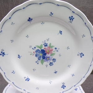 May include: White ceramic plate with a scalloped edge and a blue floral design. The plate features a central bouquet of blue, pink, and purple flowers, with smaller blue floral accents and a blue border.
