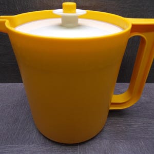 May include: A bright yellow plastic pitcher featuring a wide handle and a pouring spout. The pitcher includes a white circular lid topped with a small yellow knob handle, designed for easy removal and secure coverage of the container opening.