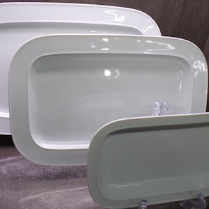 May include: A set of three white rectangular serving platters with rounded corners and a smooth, glossy finish. The platters are arranged in a cascading formation, showing a large, medium, and small size, each featuring a wide, flat rim.