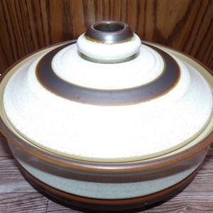 May include: A ceramic casserole dish with a lid. The dish is off-white with brown and tan stripes. The lid has a central knob and a brown ring design. The dish has two side handles. The overall design is retro.