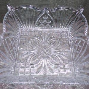 May include: Clear, square glass serving dish with a detailed floral and geometric pattern. The dish has a scalloped rim and fan-shaped designs on the sides. A decorative piece for serving snacks or displaying items.
