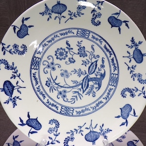 May include: White ceramic plate with a blue floral design. The plate features a central motif of a deer and floral elements, surrounded by a border of stylized flowers and fruit. The rim is decorated with additional floral and fruit patterns.