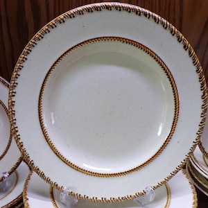 May include: A collection of off-white ceramic plates with a brown rope-like design around the rim and inner circle. The plates are stacked and displayed on clear stands, showcasing a rustic, farmhouse style.
