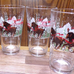 May include: Set of clear drinking glasses with a winter scene design. Each glass features a brown horse pulling a sleigh, with the words "Season's Greetings" in red script. The background includes white trees and snow.