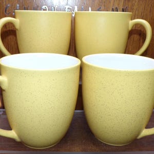 May include: Set of four yellow ceramic mugs with a speckled finish. The mugs have white interiors and rounded handles.