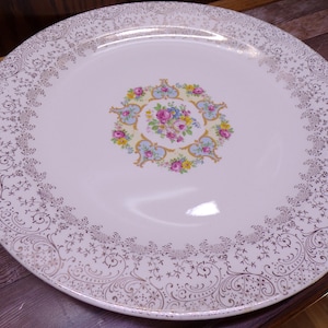 May include: A white, round vintage plate with a floral center design and gold filigree border. The center features a bouquet of pink, blue, and yellow flowers. The plate is approximately 10 inches in diameter.