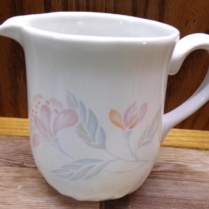 May include: White ceramic pitcher with a curved handle and a spout. The pitcher is decorated with a floral pattern in pastel pink, orange, and blue-green hues. The pitcher is designed for serving beverages.