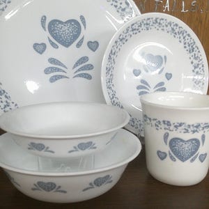 May include: A set of white Corelle dinnerware with a blue and white floral pattern featuring hearts. The set includes a large plate, a smaller plate, two bowls, and a mug.