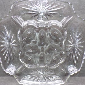 May include: Clear glass serving dish with a starburst design. The dish has a scalloped edge and a central section with a floral pattern. The starbursts are located on the corners and center of the dish.