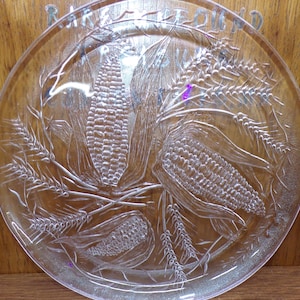 May include: A clear glass serving plate with an etched design of corn and wheat stalks. The plate has a raised rim and is perfect for serving snacks or appetizers.