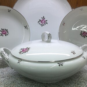 May include: A white porcelain serving dish with a lid, featuring a delicate floral design with pink roses and silver accents. The dish is displayed on a table with three matching plates.