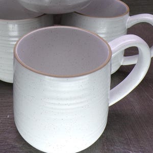 May include: A set of white ceramic mugs featuring a speckled finish and horizontal ribbed texture. Each mug has a smooth, rounded handle and a light brown rim detail. The mugs are arranged in a cluster, showcasing their cylindrical shape.