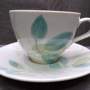 May include: A white teacup and saucer set with a leaf design in shades of green and blue. The teacup has a curved handle and sits on a matching saucer. The leaf pattern is painted on both the cup and saucer.