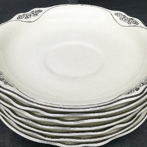 May include: A stack of eight white ceramic saucers with a black rim and floral design.