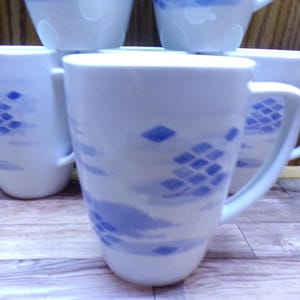 May include: White ceramic mugs with a blue abstract design. The mugs have a cylindrical shape with a square handle. The blue design features diamond and cloud-like shapes. The mugs are stacked on a wooden surface.