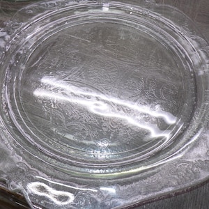 May include: Clear glass serving plate with an ornate, embossed floral and scroll design. The plate has a scalloped edge and a raised center. The glass reflects light, highlighting the intricate details of the pattern. A vintage piece for serving food.
