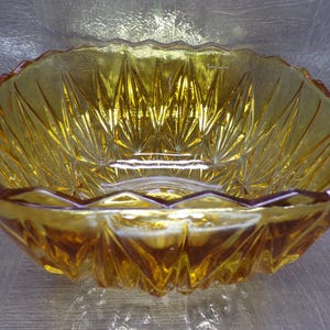 May include: A decorative amber-colored glass bowl with a scalloped rim. The bowl features a geometric, starburst-like pattern etched into the glass. The bowl is designed with a series of facets that catch and reflect light.