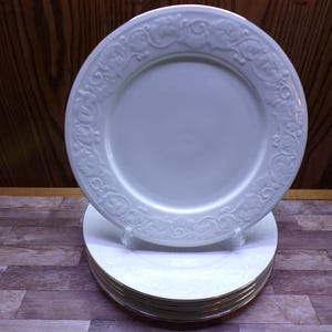 Pu&ograve; includere: Una pila di piatti in ceramica bianca con un motivo floreale in rilievo sul bordo. I piatti hanno un bordo dorato e sono esposti su una superficie di legno.