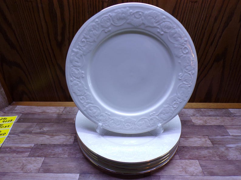 Pu&ograve; includere: Una pila di piatti in ceramica bianca con un motivo floreale in rilievo sul bordo. I piatti hanno un bordo dorato e sono esposti su una superficie di legno.