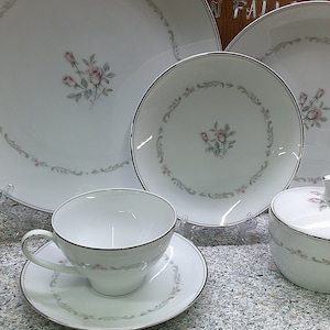 May include: A set of white porcelain dinnerware with a delicate floral design featuring pink roses and green leaves. The set includes a large dinner plate, a smaller salad plate, a teacup and saucer, and a sugar bowl with a lid.