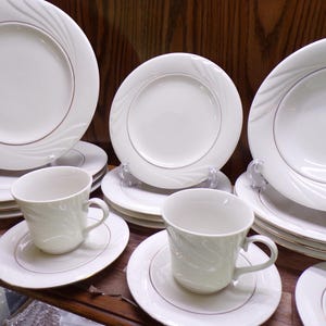 May include: A collection of white porcelain dinnerware with a gold trim. The set includes plates, bowls, saucers, and teacups. The plates and bowls have a subtle swirl design. The teacups have a matching swirl pattern.
