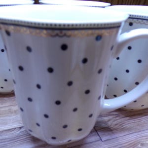May include: White ceramic mugs with black polka dots and gold trim. The mugs have a curved handle and a slightly flared shape. The mugs are arranged on a wooden surface.